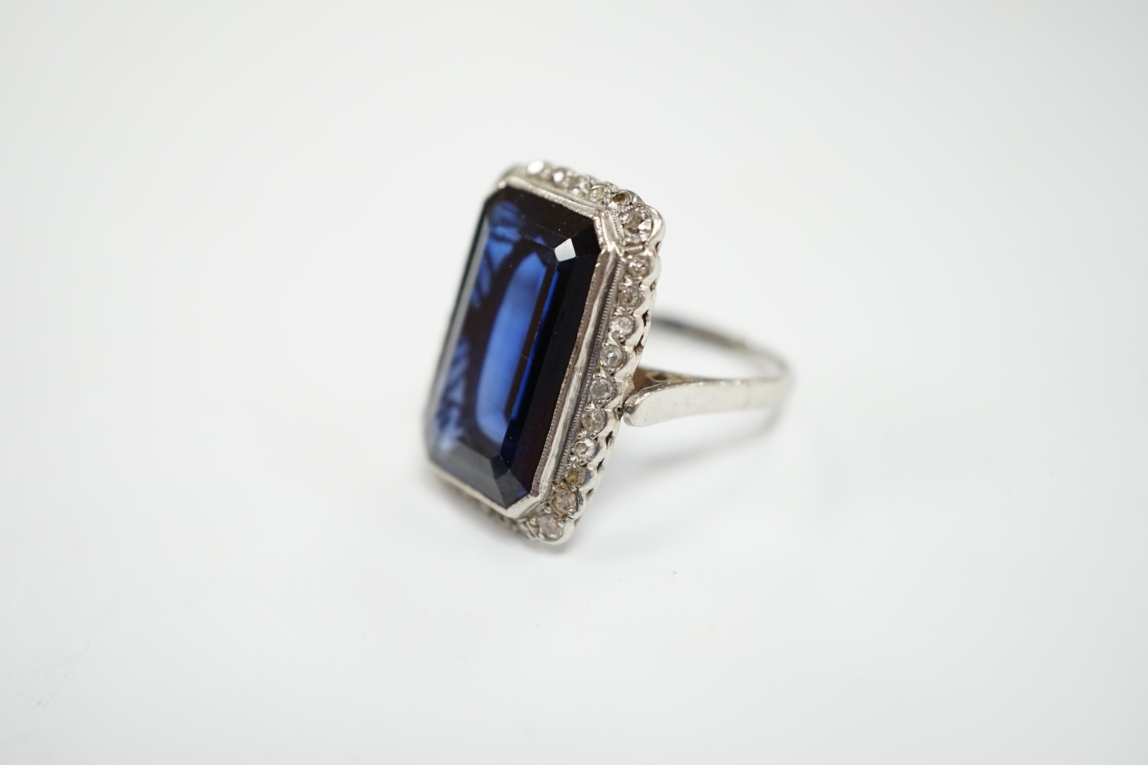A 1920's white metal and single stone emerald cut synthetic sapphire set dress ring, with millegrain set diamond chip border, size N, gross weight 10 grams.